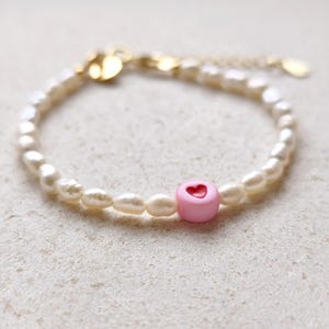 Op de afbeelding: Een delicate armband met een rij kleine, crèmekleurige parels. Een roze hartvormige kraal met een rode hartuitsparing is gecentreerd. De sluiting en ketting zijn goudkleurig.