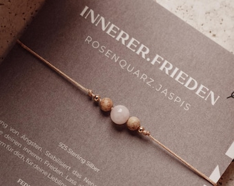 May include: A delicate bracelet featuring a central light pink bead flanked by two speckled tan beads on a thin metallic cord. The bracelet is attached to a dark gray display card with the text INNERER.FRIEDEN ROSENQUARZ.JASPIS and 925 Sterling Silber.