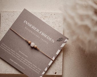 May include: A delicate bracelet featuring a central light pink rose quartz bead flanked by two tan jasper beads and small metallic accents on a thin beige cord. The bracelet is displayed on a grey card with the text INNERER.FRIEDEN ROSENQUARZ.JASPIS.