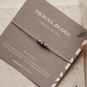 May include: A fine bracelet featuring turquoise and brown beads on a thin, beige cord. The bracelet is presented on a grey card with the text "TRAVEL.BUDDY" and "TÜRKIS.JASPIS". The card also includes text in German.