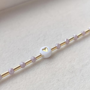 Op de afbeelding: Close-up van een delicate armband. Het heeft een witte hartvormige kraal met een gouden hartontwerp, afgewisseld met kleine, lichtpaarse kralen en goudkleurige metalen buisjes. De armband wordt gepresenteerd op een lichtgekleurd oppervlak.