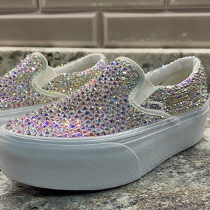 Könnte beinhalten: Weiße Slip-on-Sneaker mit Plateau-Sohle, die mit schimmernden Strasssteinen besetzt sind. Die Schuhe haben eine dicke weiße Plateau-Sohle und das Vans-Logo ist an der Seite sichtbar.