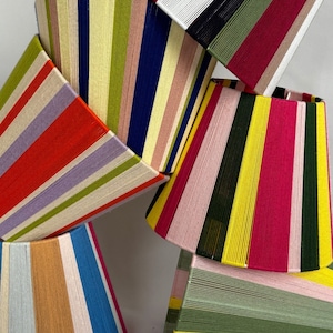 May include: A selection of lampshades with vertical stripes in a variety of colours, including red, blue, yellow, and green. The shades are stacked, displaying their unique design and vibrant colour palette. These lampshades are a decorative lighting element.