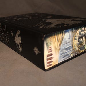 Op de afbeelding: Een zwart hardback boek met de titel "Dune Trilogy" in wit op de rug. De omslag toont een gedetailleerde illustratie van een landschap met een stel op de voorgrond.
