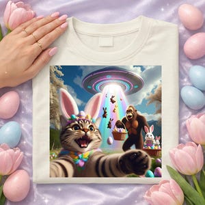 May include: Off-white t-shirt featuring a colorful graphic of a cat wearing bunny ears, taking a selfie with a gorilla, an alien spaceship, and Easter eggs. The cat has a colorful floral collar. The background includes a rainbow and Easter bunnies.