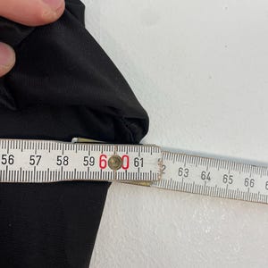 Op de afbeelding: Een zwart kledingstuk wordt gemeten met een houten liniaal. De meting is ongeveer 60 cm. Het kledingstuk is gemaakt van een gladde, donkere stof. De achtergrond is een effen wit oppervlak.