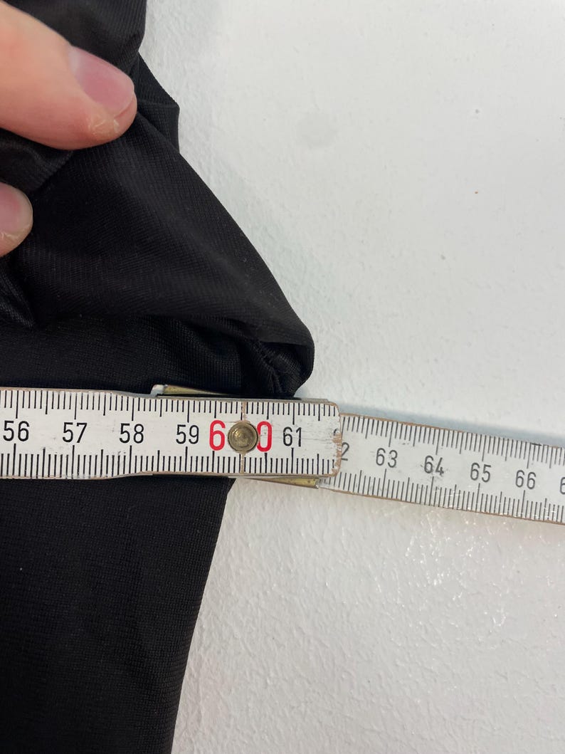 Op de afbeelding: Een zwart kledingstuk wordt gemeten met een houten liniaal. De meting is ongeveer 60 cm. Het kledingstuk is gemaakt van een gladde, donkere stof. De achtergrond is een effen wit oppervlak.
