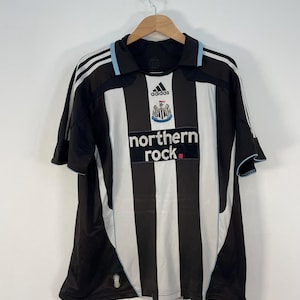 Op de afbeelding: Zwart-wit gestreept voetbalshirt met de tekst "northern rock" en het Adidas-logo. Het shirt heeft een lichtblauwe bies en hangt aan een houten hanger.