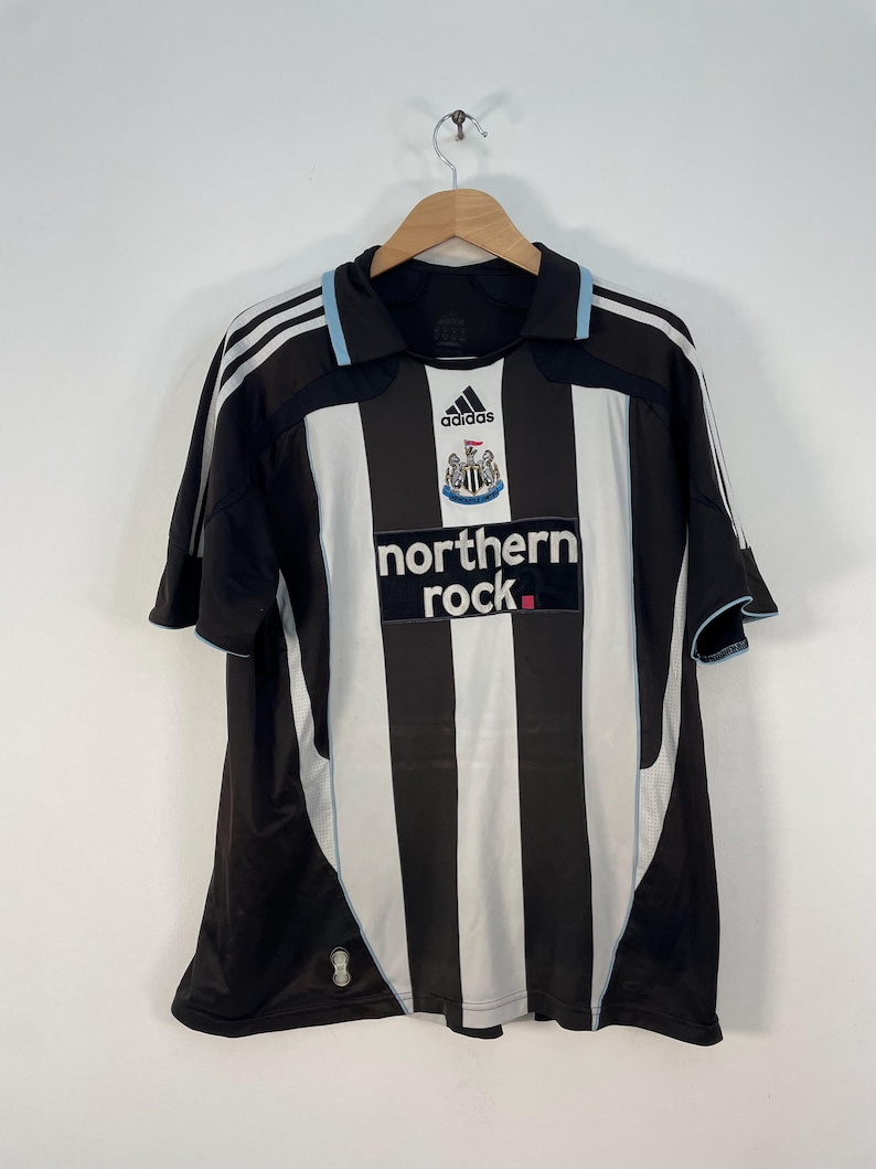Op de afbeelding: Zwart-wit gestreept voetbalshirt met de tekst "northern rock" en het Adidas-logo. Het shirt heeft een lichtblauwe bies en hangt aan een houten hanger.