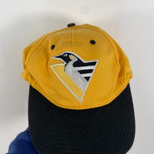Könnte beinhalten: Eine gelbe und schwarze Baseballkappe mit einem Pinguin-Logo. Das Logo ist ein schwarz-weißer Pinguin mit einem gelben Schnabel und einem gelb-schwarz gestreiften Muster auf der Brust. Das Logo befindet sich auf einem gelben Dreieck mit schwarzem Umriss.