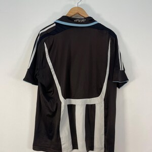 Op de afbeelding: Een bruin en zwart voetbalshirt met witte en lichtblauwe accenten. Het shirt heeft witte strepen aan de zijkanten en een lichtblauwe kraag. De tekst "NUFC" is zichtbaar op de kraag.