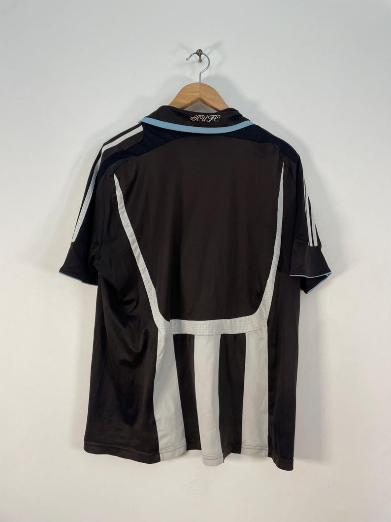 Op de afbeelding: Een bruin en zwart voetbalshirt met witte en lichtblauwe accenten. Het shirt heeft witte strepen aan de zijkanten en een lichtblauwe kraag. De tekst "NUFC" is zichtbaar op de kraag.