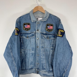 May include: Light blue denim jacket with button closure and two front pockets. Features embroidered patches with the American flag and the words "USA" and "ARMY RETIRED". The jacket is hanging on a wooden hanger.