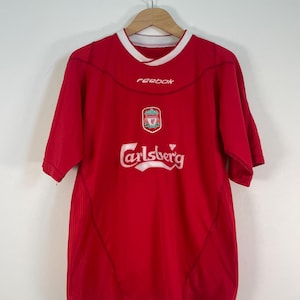 May include: Red short-sleeved jersey with white trim and the Reebok logo. The jersey features the Liverpool crest and the Carlsberg logo. The jersey is hanging on a wooden hanger against a white background.