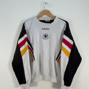 Könnte beinhalten: Weißer Adidas-Sweatshirt mit schwarz-rot gestreiften Ärmeln. Der Sweatshirt trägt das Adidas-Logo und ein Wappen der deutschen Nationalmannschaft auf der Vorderseite.