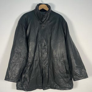 Könnte beinhalten: Schwarze Lederjacke mit hohem Kragen und Knopfverschluss, an einem Holzbügel hängend. Die Jacke hat zwei Vordertaschen und ein leicht abgenutztes Aussehen, was auf einen Vintage-Stil hindeutet.