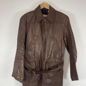 May include: A brown leather jacket with a collared neckline, button closure, and a belt. The jacket has two front pockets and is hanging on a wooden hanger. The leather has a textured appearance.