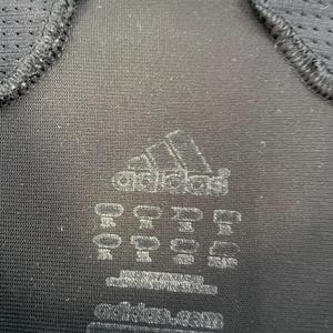 Op de afbeelding: Close-up van een zwart sportshirt met het Adidas-logo en diverse labeldetails. Het shirt heeft mesh panelen bij de schouders en halslijn. Het Adidas-logo is wit.