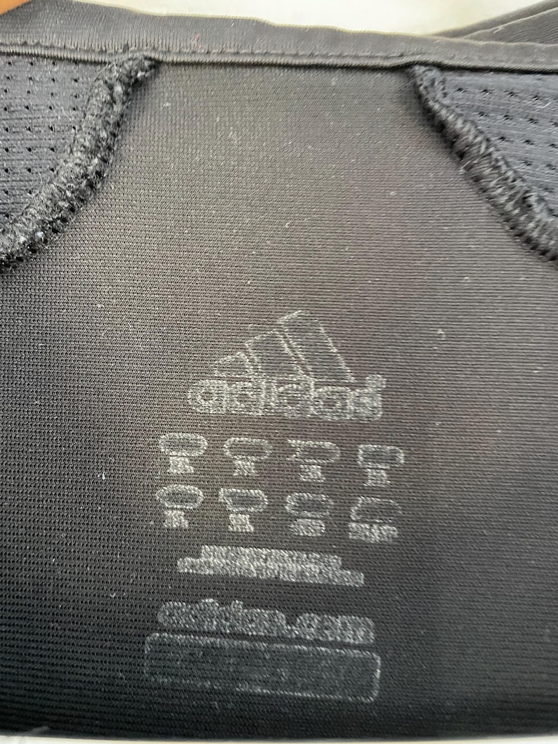 Op de afbeelding: Close-up van een zwart sportshirt met het Adidas-logo en diverse labeldetails. Het shirt heeft mesh panelen bij de schouders en halslijn. Het Adidas-logo is wit.