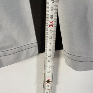 Op de afbeelding: Close-up van een grijze en zwarte stof met een meetlint. Het meetlint toont metingen van 165 tot 221 cm. De stof heeft een verticale zwarte streep in het midden.
