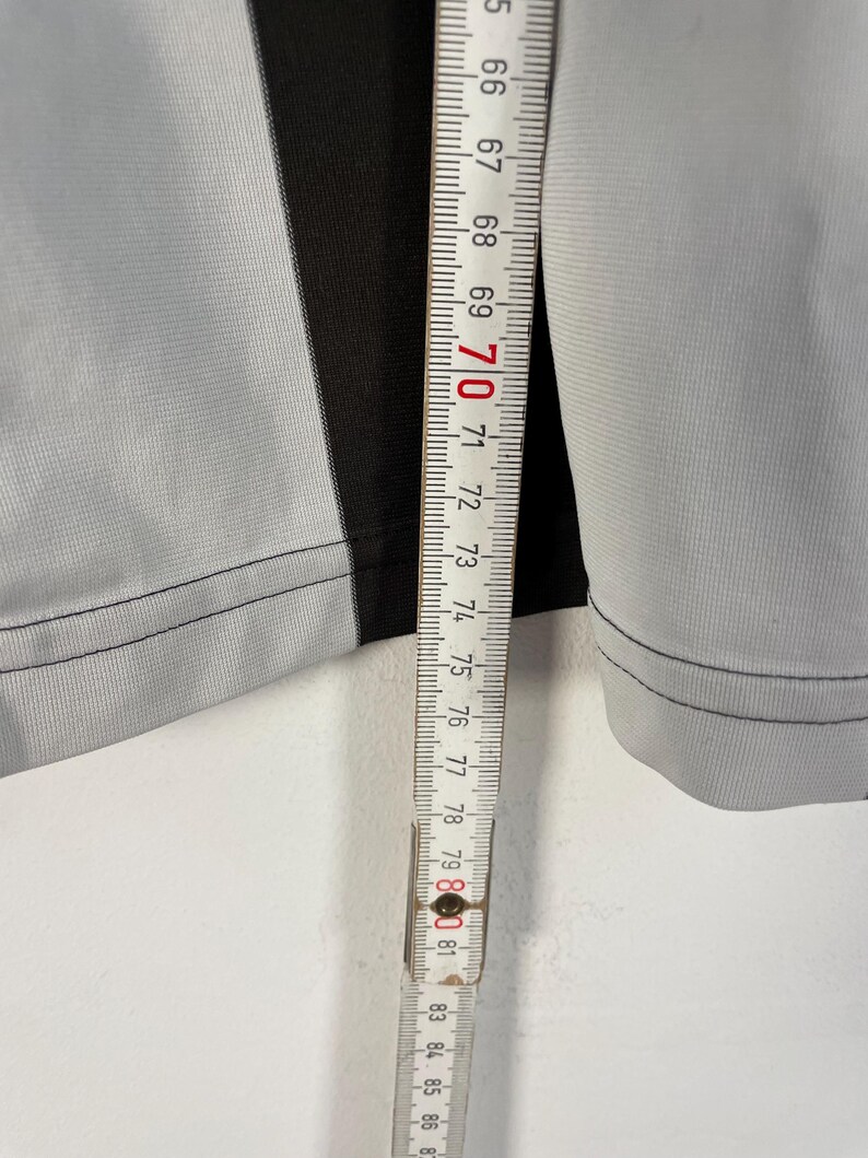 Op de afbeelding: Close-up van een grijze en zwarte stof met een meetlint. Het meetlint toont metingen van 165 tot 221 cm. De stof heeft een verticale zwarte streep in het midden.