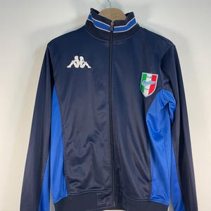 Op de afbeelding: Marineblau sportjack met een volledige ritssluiting, blauwe zijpanelen en een wit Kappa-logo. De jas heeft een geribbelde kraag en manchetten met blauw-witte strepen en een teamembleem. Klassieke sportkleding.