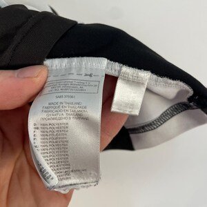 Op de afbeelding: Close-up van een zwart-wit kledinglabel. Het label bevat tekst in meerdere talen, waaronder Engels, Frans en Spaans, en geeft aan dat het materiaal 100% polyester is. Het label is bevestigd aan een zwart kledingstuk.
