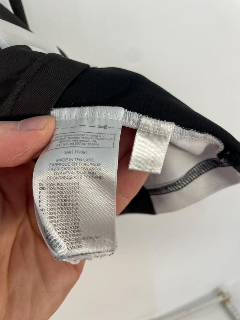 Op de afbeelding: Close-up van een zwart-wit kledinglabel. Het label bevat tekst in meerdere talen, waaronder Engels, Frans en Spaans, en geeft aan dat het materiaal 100% polyester is. Het label is bevestigd aan een zwart kledingstuk.