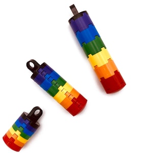 Könnte beinhalten: Drei regenbogenfarbene, zylindrische Flaschenverschlüsse mit schwarzen Schlaufen. Jeder Verschluss besteht aus gestapelten, ineinandergreifenden Segmenten in einem Farbspektrum: Rot, Orange, Gelb, Grün, Blau und Lila. Die Verschlüsse sind diagonal vor weißem Hintergrund angeordnet.