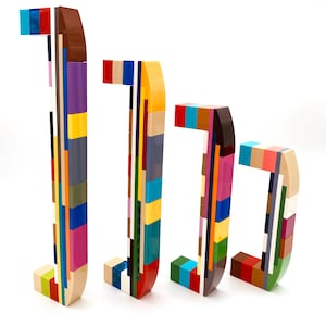 Könnte beinhalten: Vier farbenfrohe, gestapelte Buchstaben J-Skulpturen aus verschiedenfarbigen Blöcken. Die Buchstaben sind in einer Reihe angeordnet, jede in einer anderen Größe, mit einem weißen Hintergrund. Die Blöcke sind in Rot-, Blau-, Gelb-, Braun- und Grüntönen.