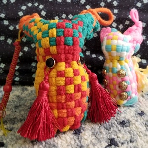 May include: Two colorful woven fabric toys. One is a red, yellow, and green woven pouch with red tassels. The other is a pink, white, and yellow woven pouch with gold buttons.