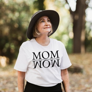 Puede incluir: Una mujer con una camiseta blanca con el texto "MOM WOW" impreso en ella. Lleva un sombrero gris y mira hacia arriba.