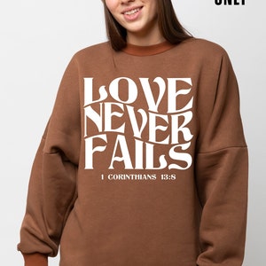 Puede incluir: Una sudadera marrón con el texto "LOVE NEVER FAILS" en blanco, con la referencia bíblica "1 CORINTHIANS 13:8" debajo del texto.
