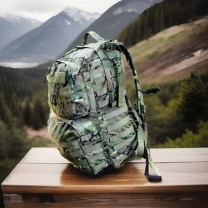 Könnte beinhalten: Ein grüner und brauner Tarnrucksack mit mehreren Riemen und Taschen. Der Rucksack steht auf einer Holzoberfläche.