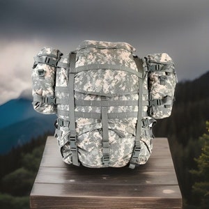 Puede incluir: Una mochila grande de camuflaje verde y marrón con múltiples correas y hebillas. La mochila está sentada sobre una superficie de madera.