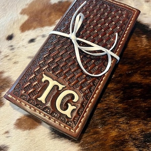May include: A brown leather wallet with a woven pattern and the initials "TG" in a lighter color. The wallet is secured with a white ribbon. The background is a cowhide pattern.