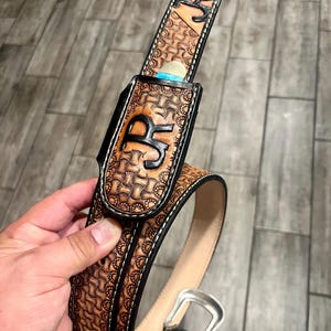 May include: A handcrafted leather belt with a decorative pattern and the initials "JR" embossed on the holster. The belt features a silver buckle and a contrasting black border with white stitching. The holster appears to be designed to hold a small item.