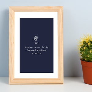 Puede incluir: Una impresión enmarcada con un fondo azul marino y texto blanco que dice "You're never fully dressed without a smile". Una pequeña ilustración de micrófono está por encima del texto. El marco es de madera clara y está sobre una superficie blanca junto a un pequeño cactus en maceta.