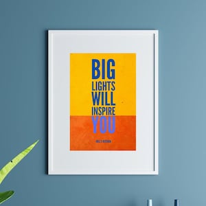 May include: A framed art print with the text "BIG LIGHTS WILL INSPIRE YOU" in blue and white on a yellow and orange background. The print hangs on a blue wall above a blue sofa and a green plant.