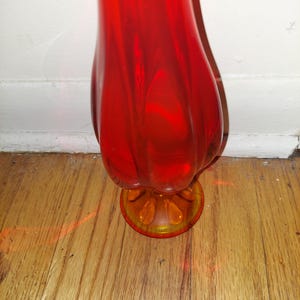 May include: A tall, red glass vase with a flared, rounded body and a smaller, round base. The base is a lighter shade of orange.