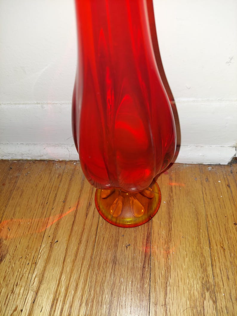 May include: A tall, red glass vase with a flared, rounded body and a smaller, round base. The base is a lighter shade of orange.