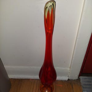 May include: A tall, red glass vase with a swirl design in yellow and orange. The vase has a rounded base and a narrow neck.
