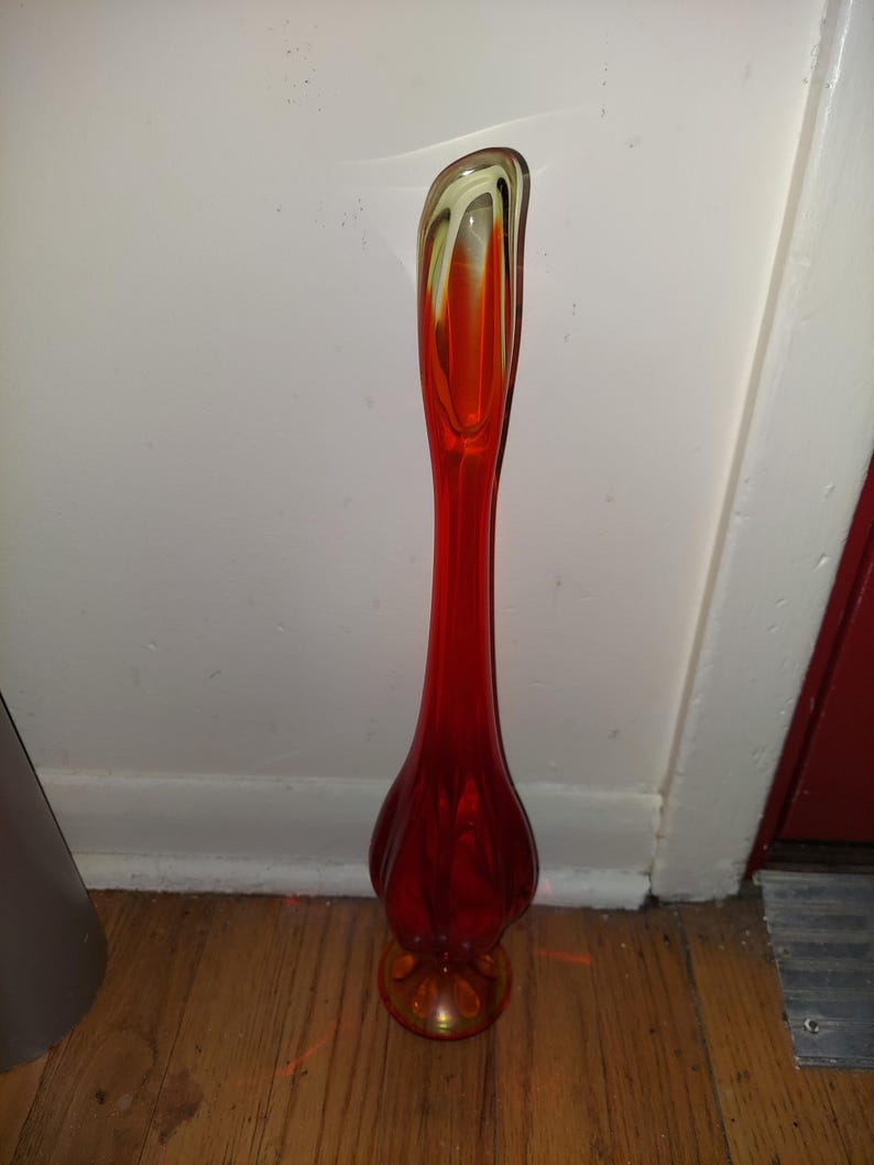 May include: A tall, red glass vase with a swirl design in yellow and orange. The vase has a rounded base and a narrow neck.
