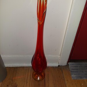 May include: A tall, red glass vase with a flared, rounded base and a thin, elongated neck. The vase has a yellow-tinted glass rim.