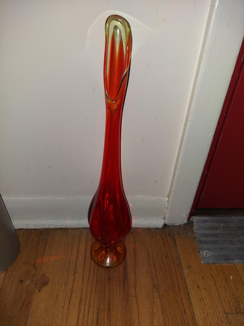 May include: A tall, red glass vase with a flared, rounded base and a thin, elongated neck. The vase has a yellow-tinted glass rim.