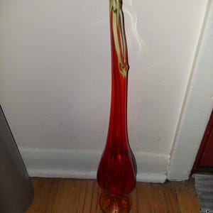 May include: A tall, red glass vase with a yellow-tinted base. The vase has a unique, curved shape and a slender neck.