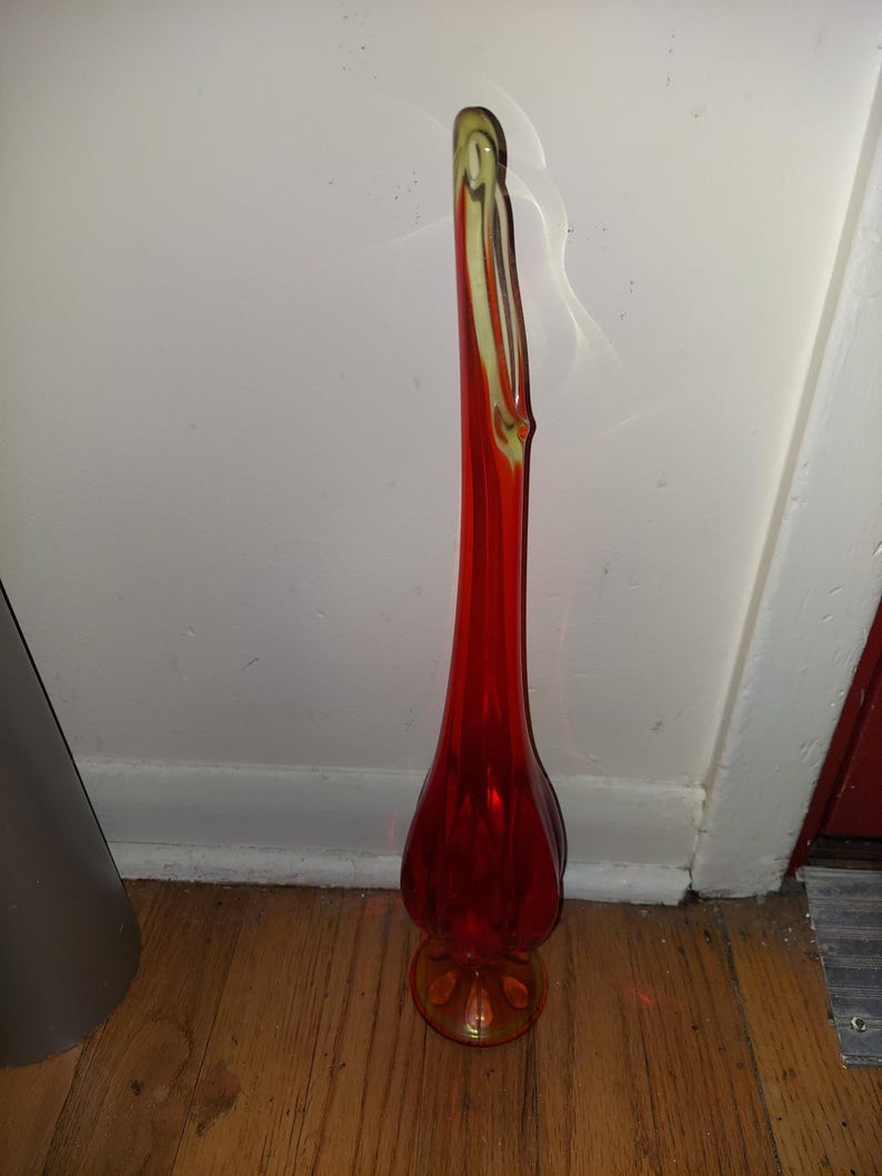 May include: A tall, red glass vase with a yellow-tinted base. The vase has a unique, curved shape and a slender neck.