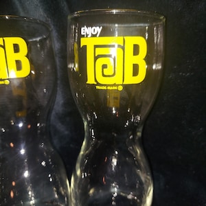 Puede incluir: Dos vasos de vidrio transparente con forma de reloj de arena para beber con un logotipo amarillo "Enjoy Tab" impreso en la parte delantera. El logotipo está en un estilo de fuente retro.
