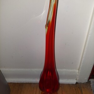 May include: A tall, red glass vase with a flared base and a twisted neck. The vase is made of clear glass with a red and orange swirl.