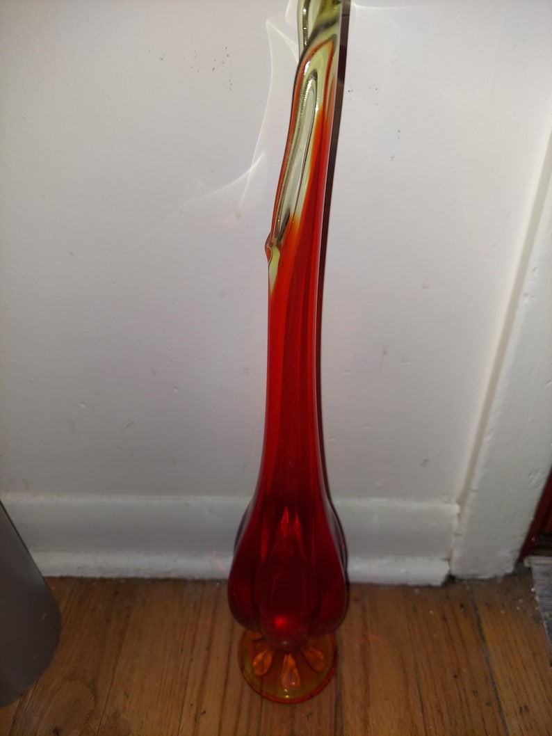 May include: A tall, red glass vase with a flared base and a twisted neck. The vase is made of clear glass with a red and orange swirl.